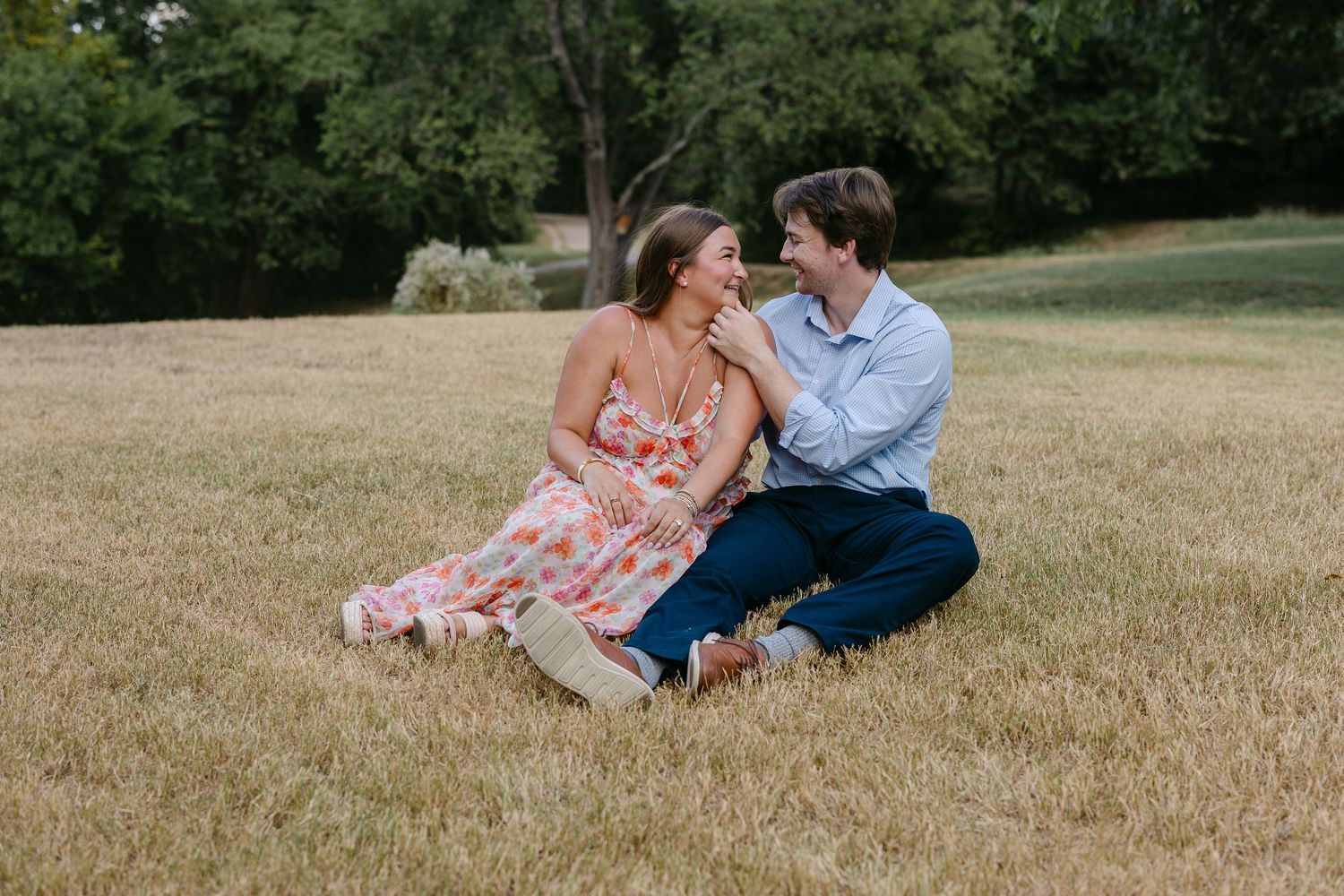 Couple sitting together on green grass field, woman in floral dress and man in blue shirt, smiling at each other outdoors.