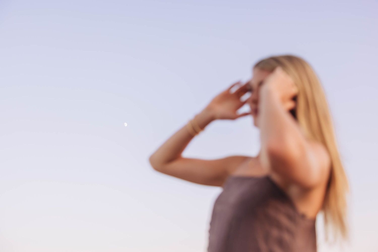 Blurred portrait of a blonde woman shielding her eyes against a clear sky outdoors.