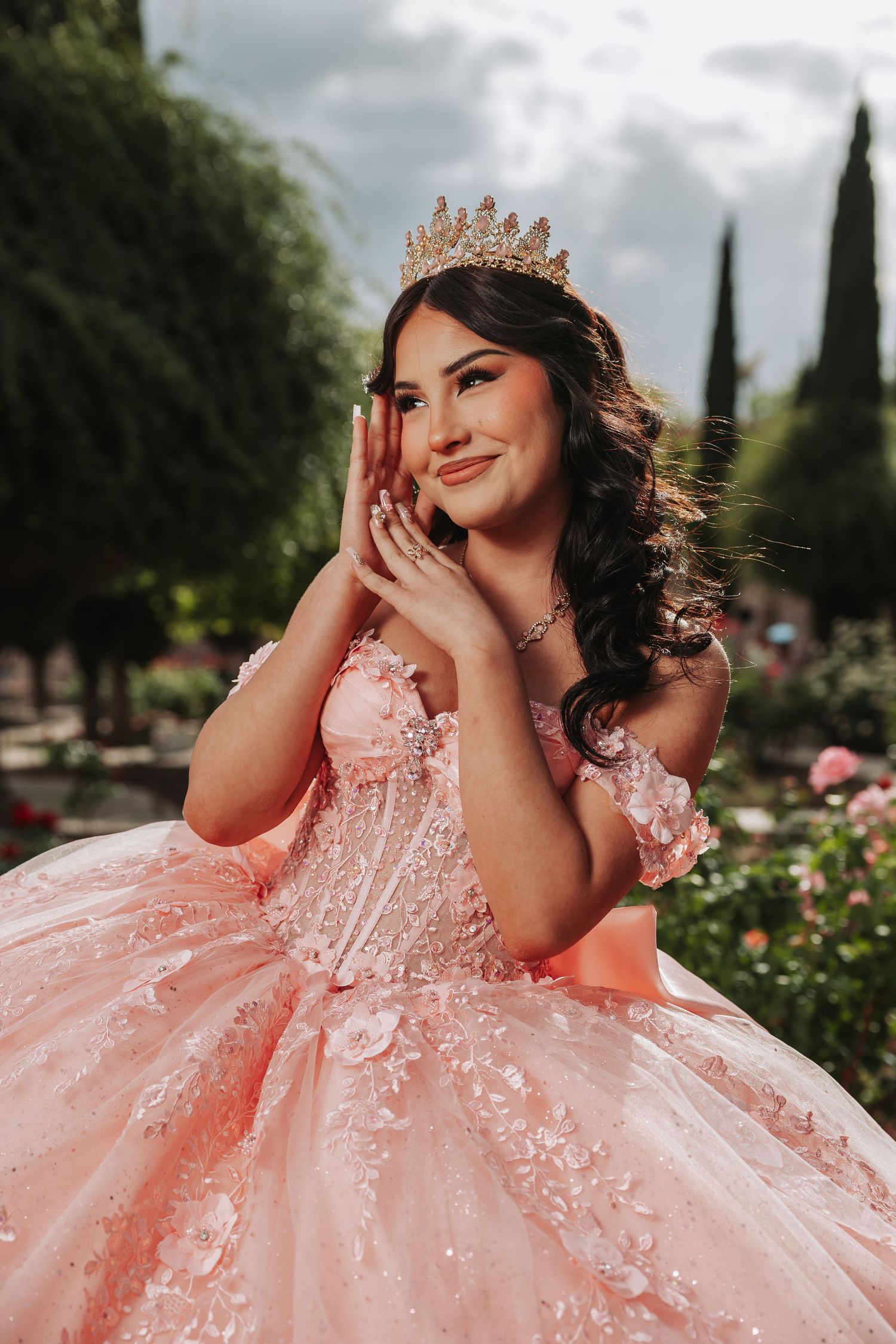 Young woman in pink quinceañera ballgown and gold crown poses gracefully in a garden setting.