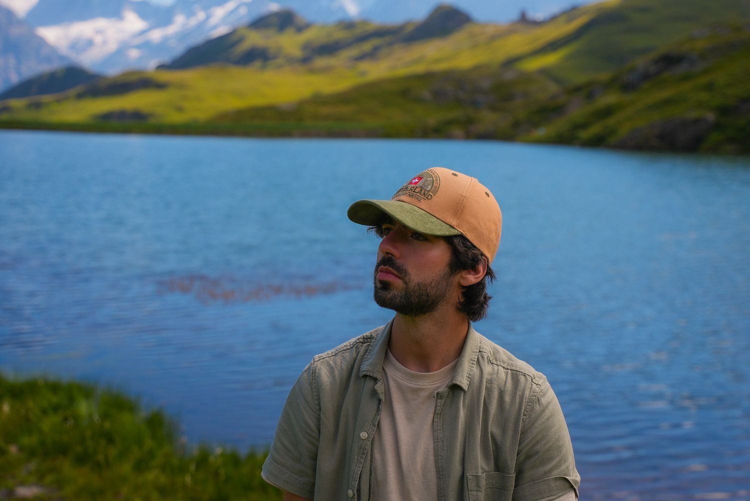 Man in tan cap and gray shirt sitting by a serene alpine lake with green hills and snow-capped mountains.
