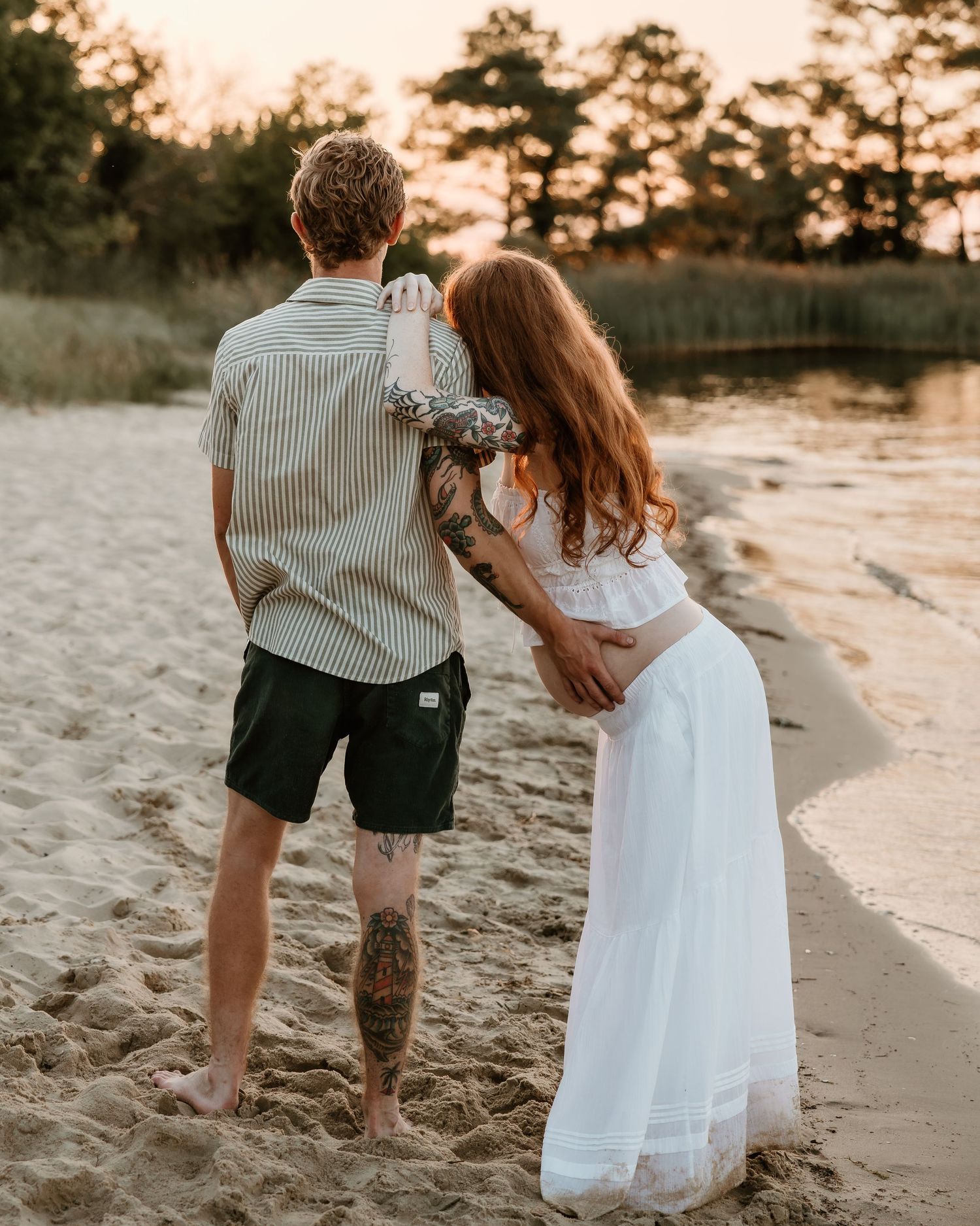 Couple holding hands on sandy beach at sunset, woman in white dress and man in striped shirt with tattoos.