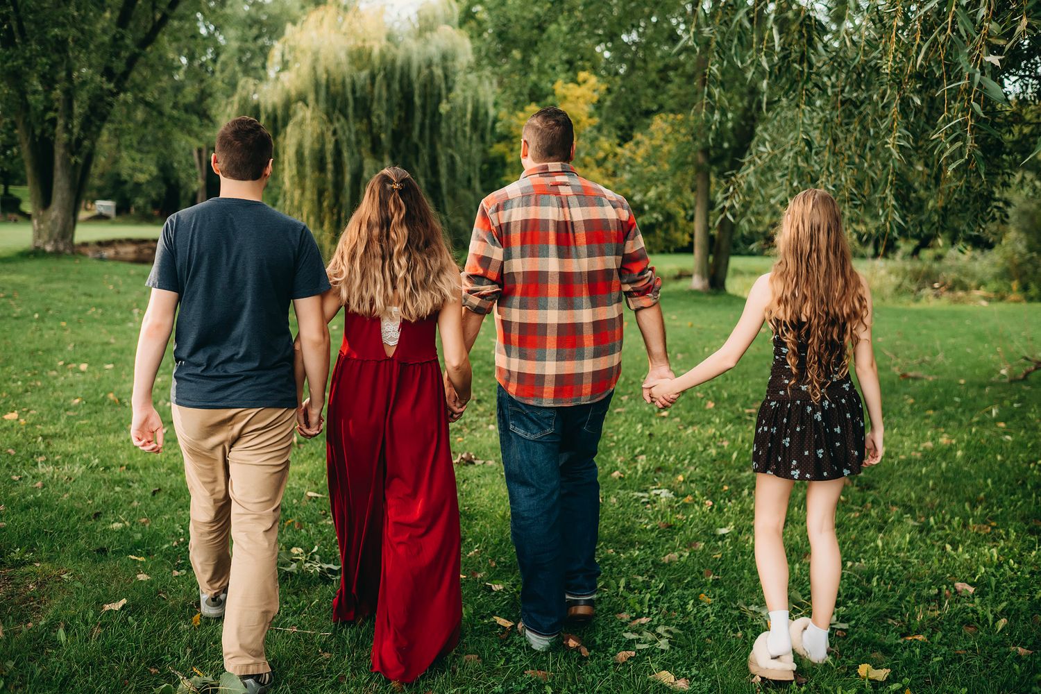 Family of four walking hand-in-hand through a lush green park, viewed from behind on a warm autumn day.