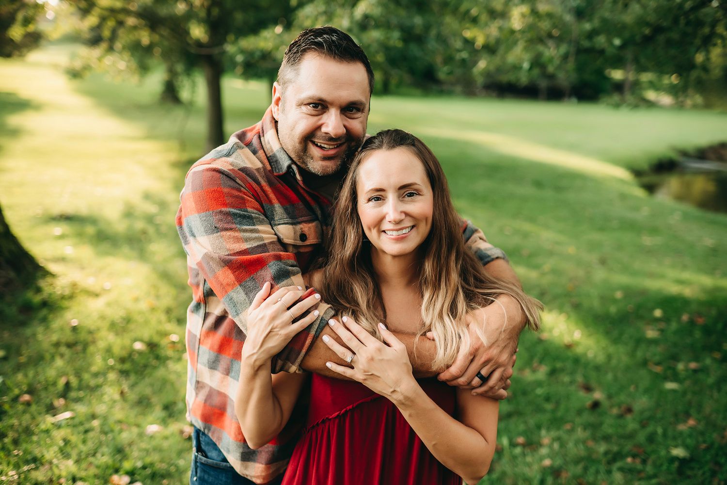 Happy couple embracing outdoors in a lush green park, woman wearing red dress, man in plaid shirt, smiling warmly.