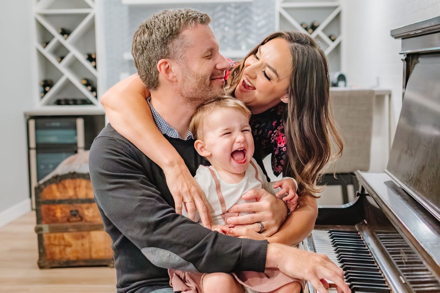 Happy family with parents kissing laughing baby sitting at piano in bright living room.