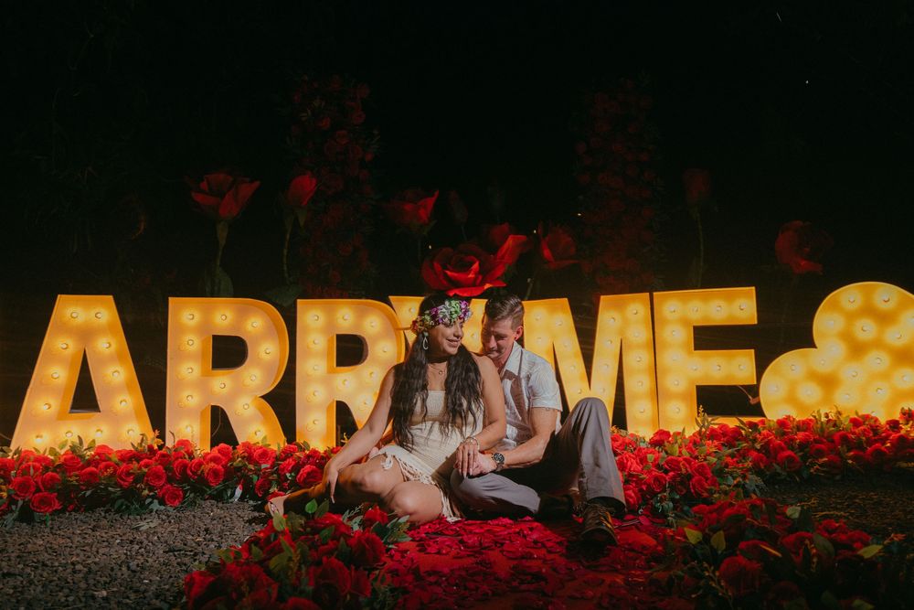 Couple sits romantically among red roses in front of illuminated marquee letters at night.