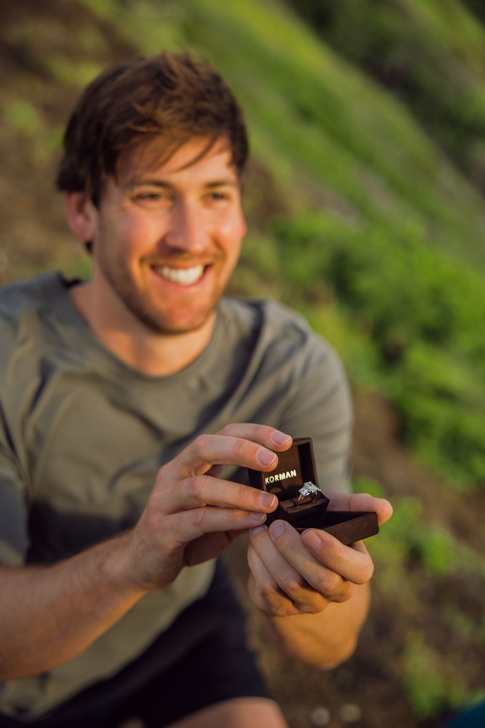 Smiling man holding open ring box outdoors, preparing to propose marriage with a diamond engagement ring.