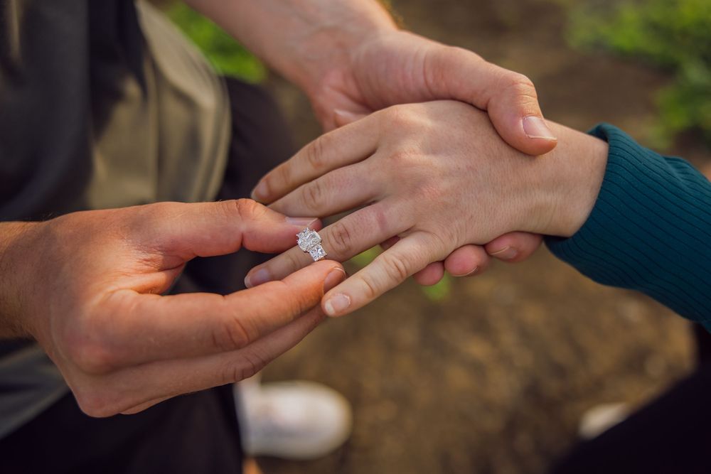 Person placing a diamond engagement ring on their partner's finger during a romantic outdoor marriage proposal.