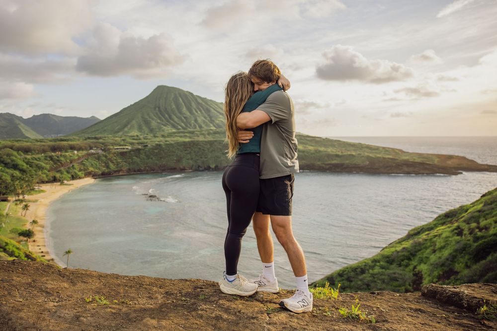Couple embracing on a scenic overlook above a tropical bay with lush green mountains and sandy beach in Hawaii.