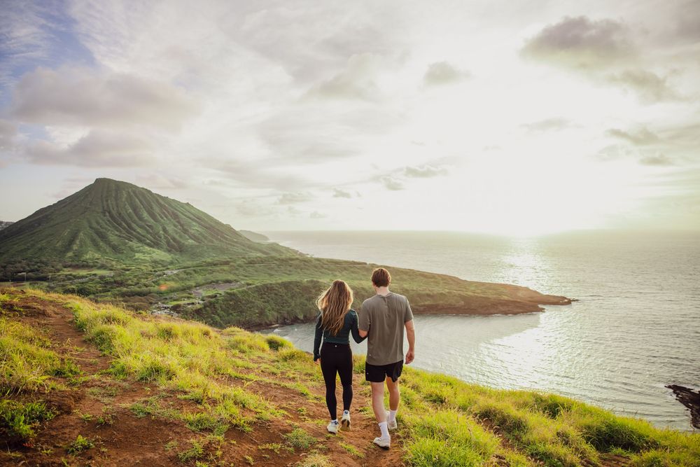 Couple holding hands walking along a scenic coastal hiking trail with lush green hills and ocean views.