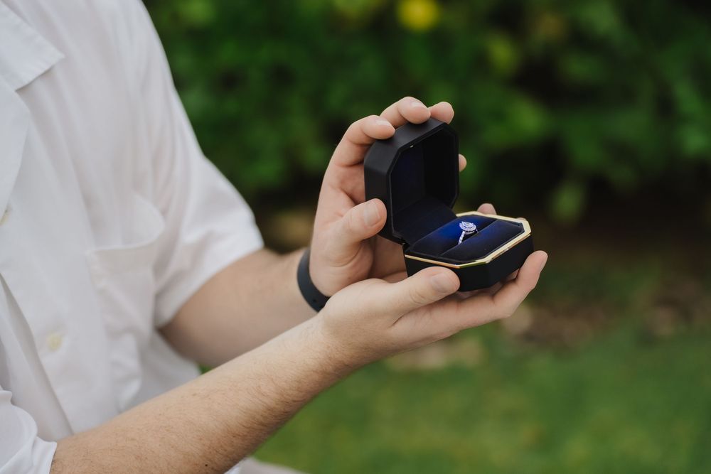 Man holding open black velvet ring box with diamond engagement ring outdoors in garden setting.