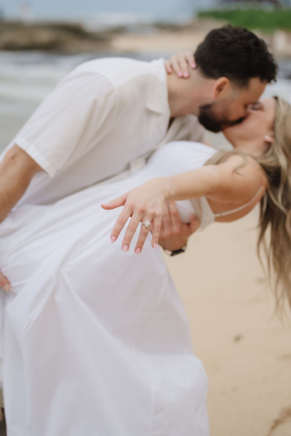 Couple kissing on beach in white outfits, woman displaying diamond engagement ring while being dipped by her partner.