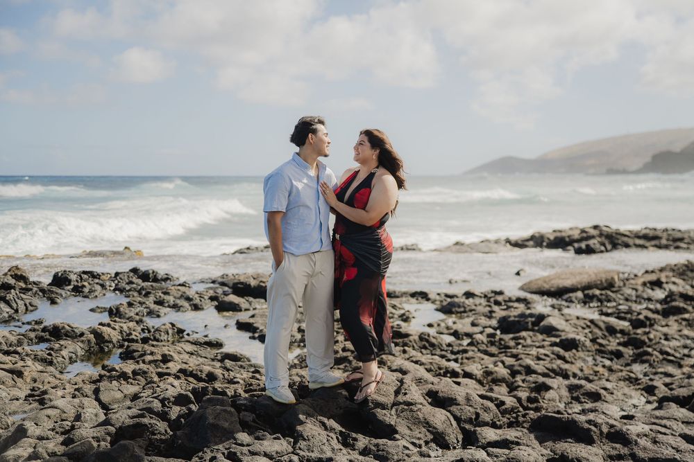 Couple standing on rocky shoreline with ocean waves behind them, sharing a romantic moment during a beach engagement photo session.