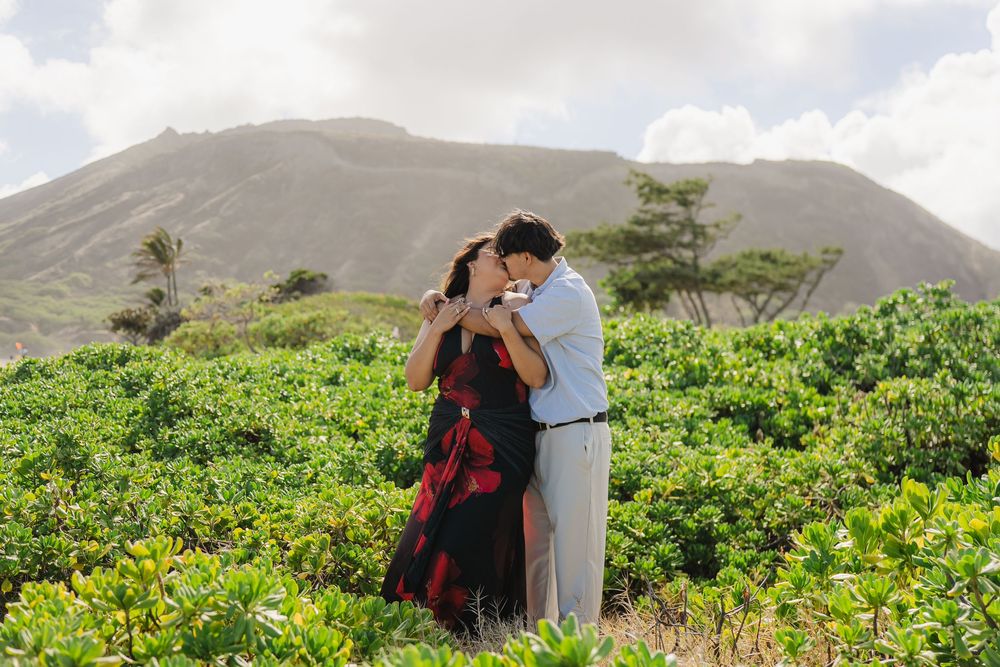 Couple embracing and kissing in a lush green field with a volcanic mountain backdrop in Hawaii.
