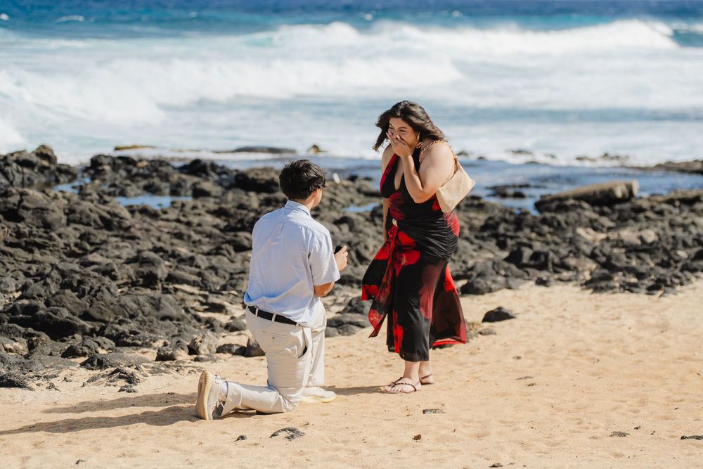 Man proposes on bended knee to woman in red dress on sandy beach with rocky shoreline and ocean waves.