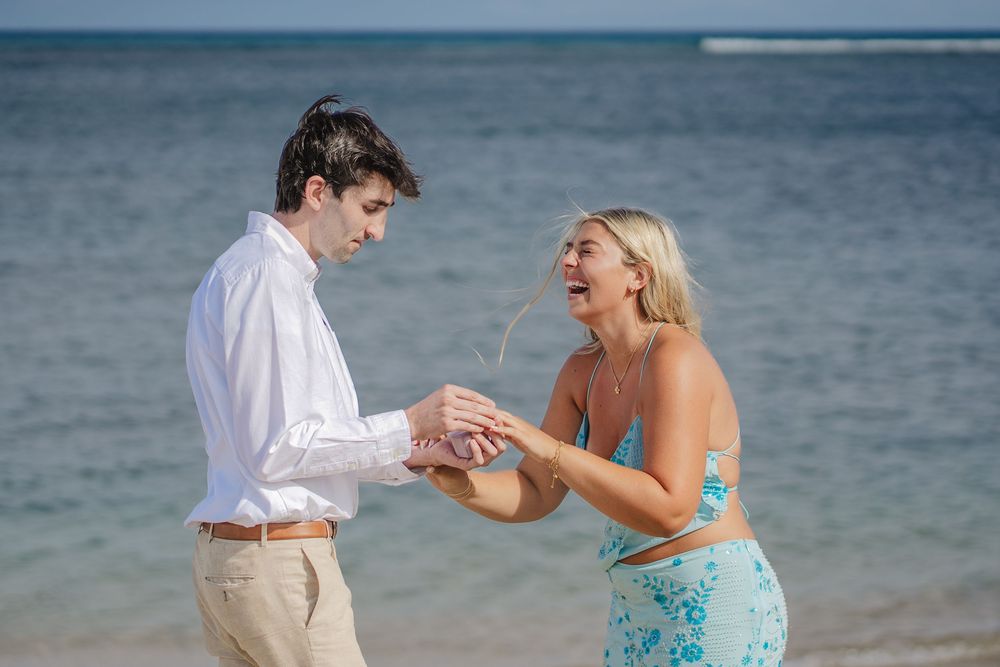 Couple sharing a joyful moment on a tropical beach, man in white shirt placing ring on laughing blonde woman.