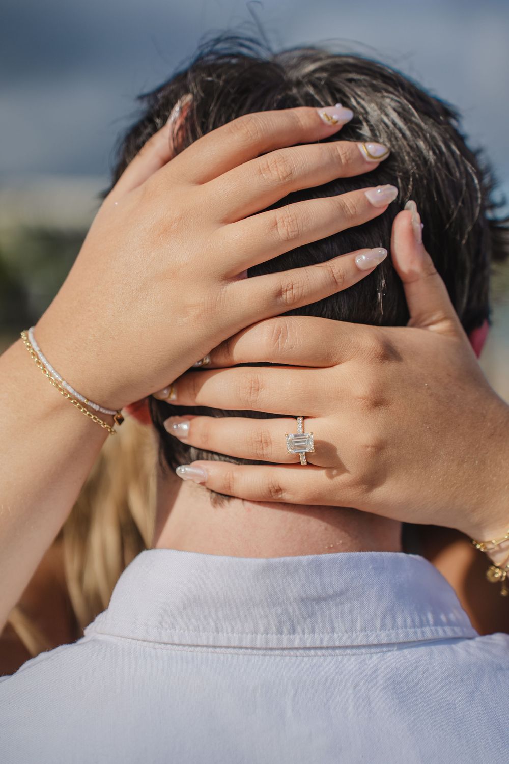Close-up of a woman's hands with an emerald-cut engagement ring and gold bracelet resting on a man's head.
