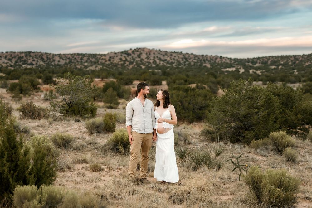 Couple stands together in desert landscape wearing white dress and khakis with shrubs and hills at sunset.