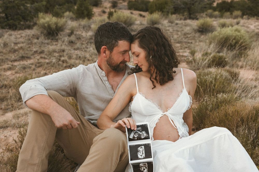 Couple sitting outdoors holding ultrasound photos during maternity shoot in desert landscape.