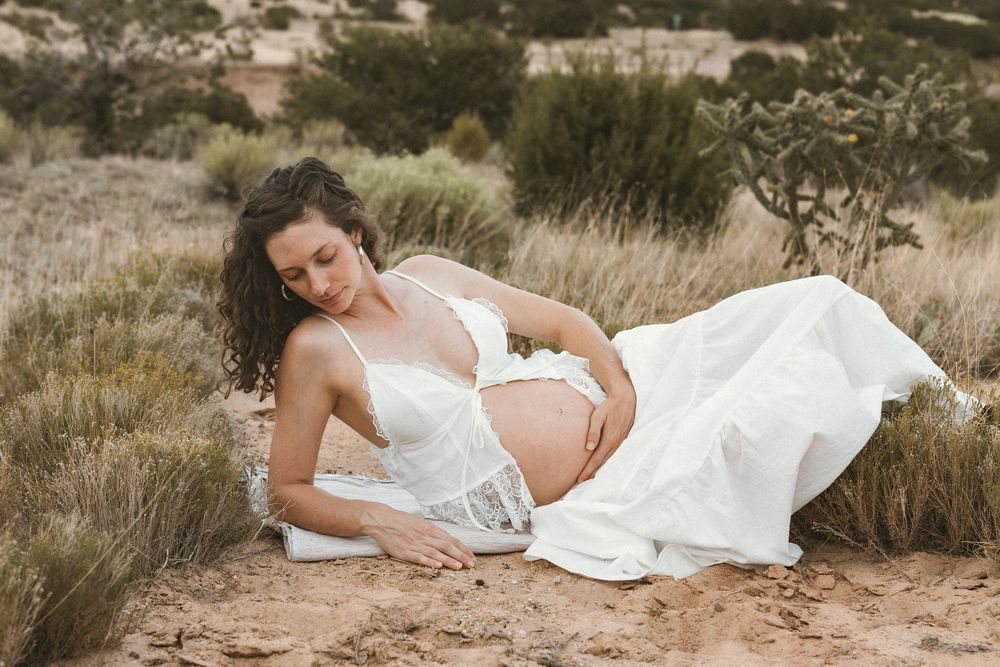 Pregnant woman in white lace dress sitting in desert landscape, cradling baby bump during outdoor maternity photoshoot.
