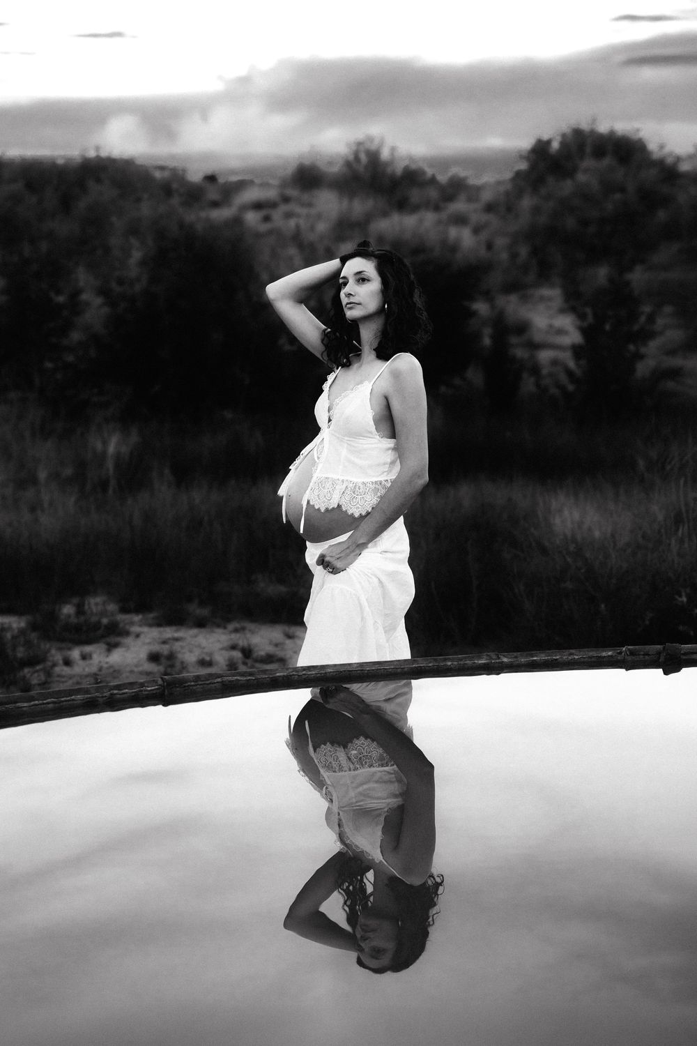 Black and white photo of pregnant woman in white dress standing by reflective pool with lush greenery backdrop.