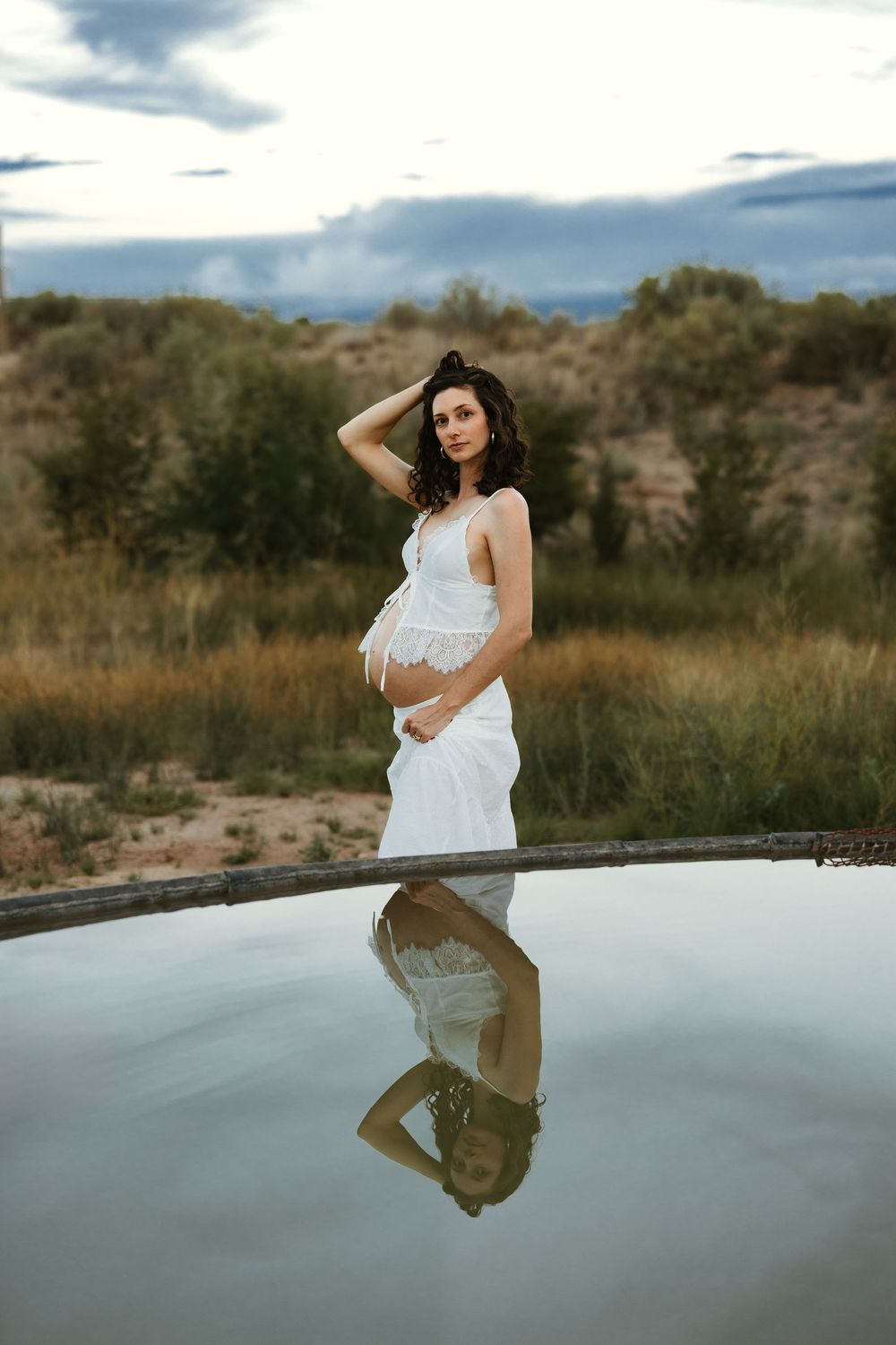 Pregnant woman in white dress stands by reflective pool in nature, cradling belly with hand in hair under cloudy sky.