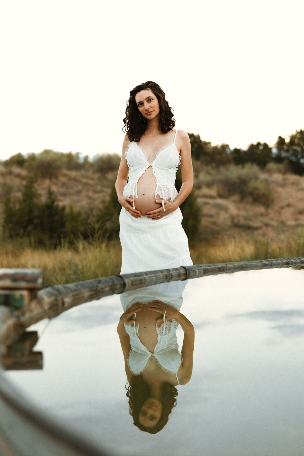 Pregnant woman in white dress holds baby bump outdoors near reflective pool with natural landscape backdrop.