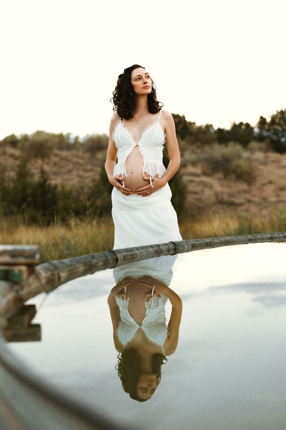Pregnant woman in white dress stands by reflective pool in nature, cradling her baby bump at golden hour.