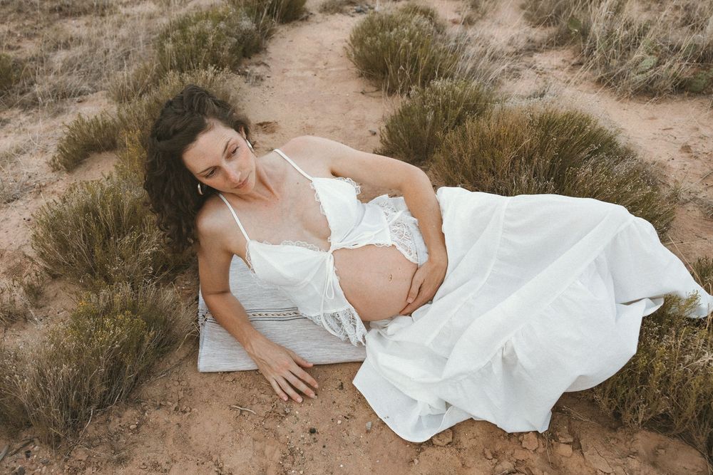 Pregnant woman in white dress lying on sandy ground outdoors, cradling her baby bump during a maternity photo shoot.