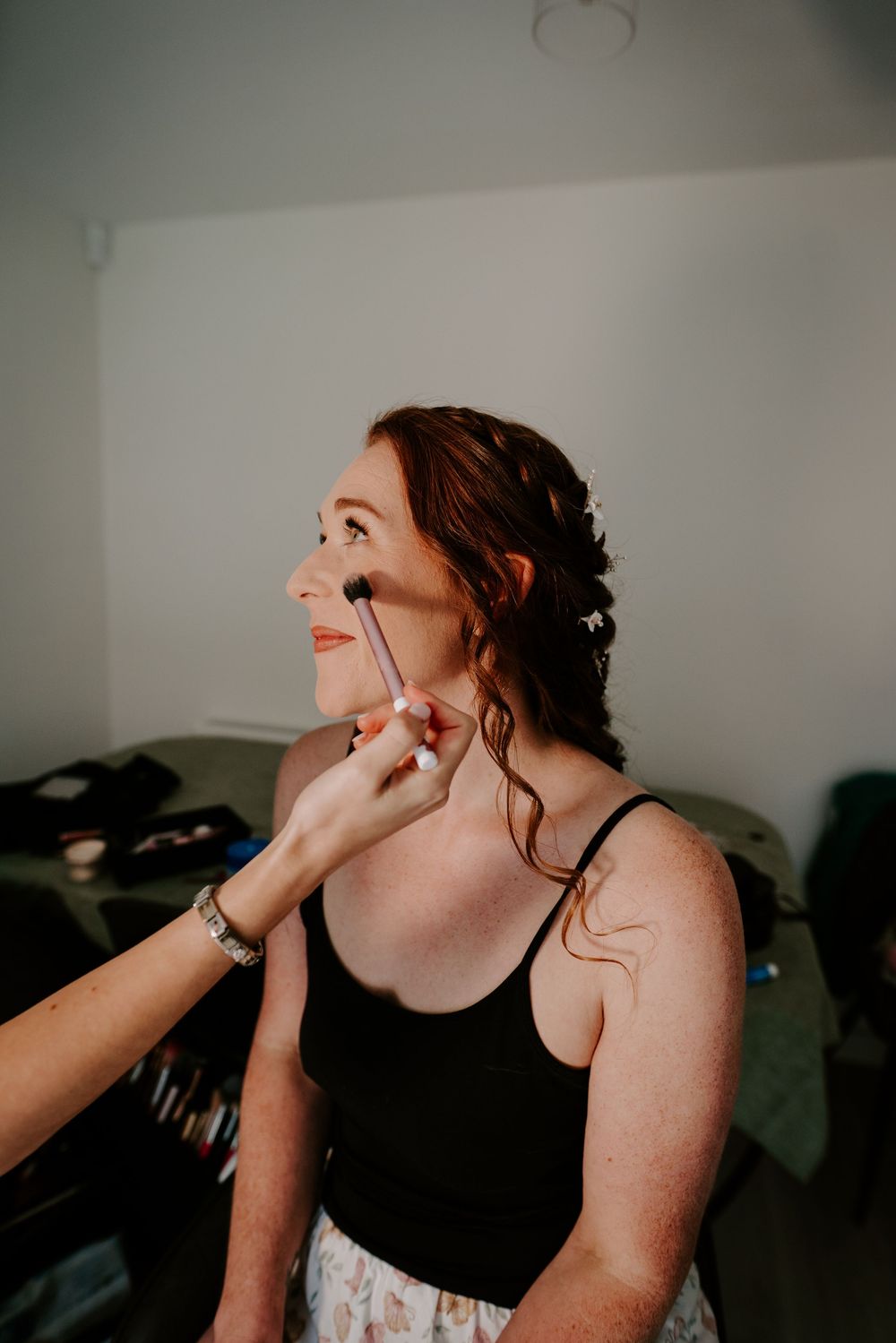Bride getting makeup applied with a brush while smiling during wedding preparations indoors.