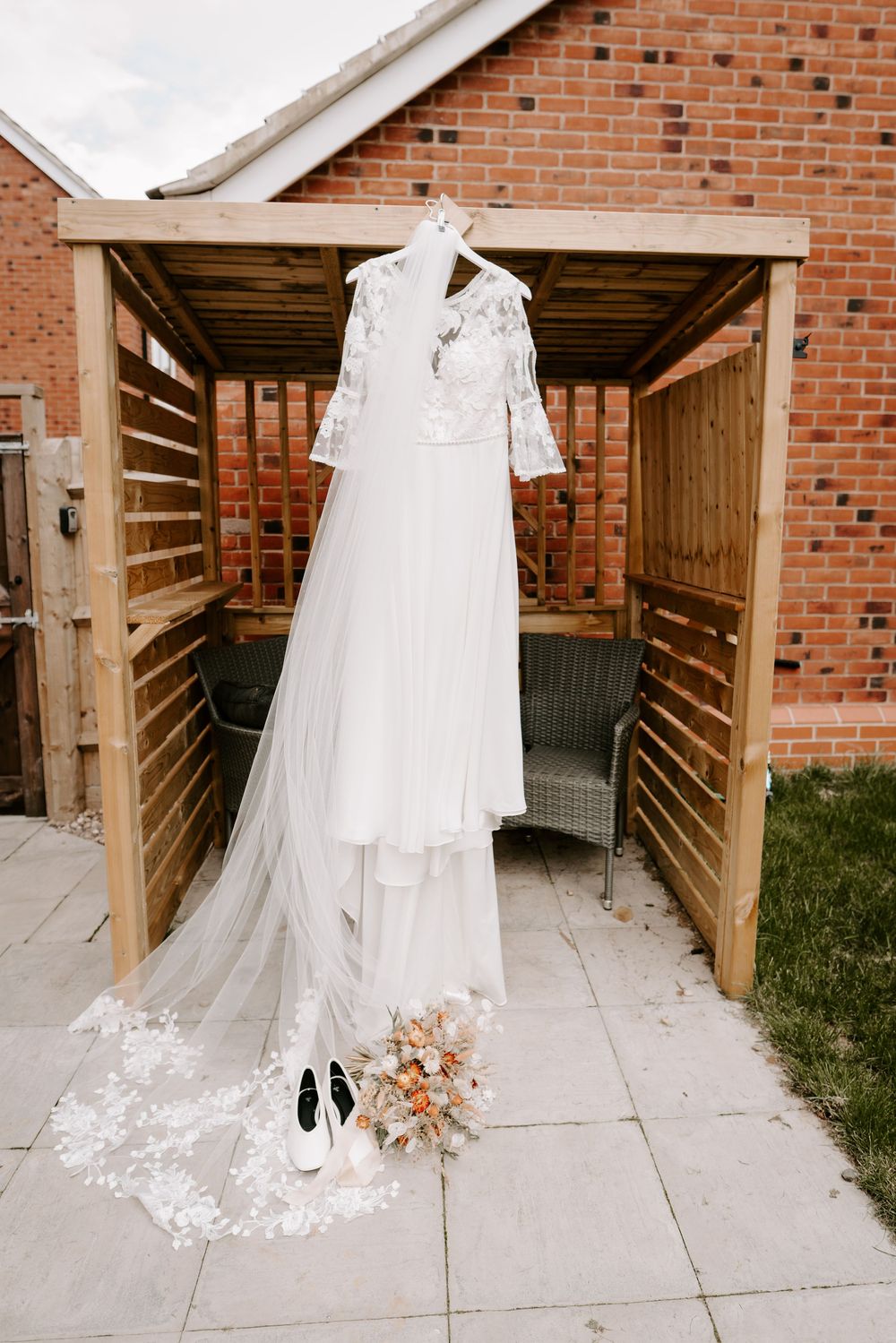 White lace wedding dress with veil hanging on wooden pergola, with bridal bouquet and shoes below.