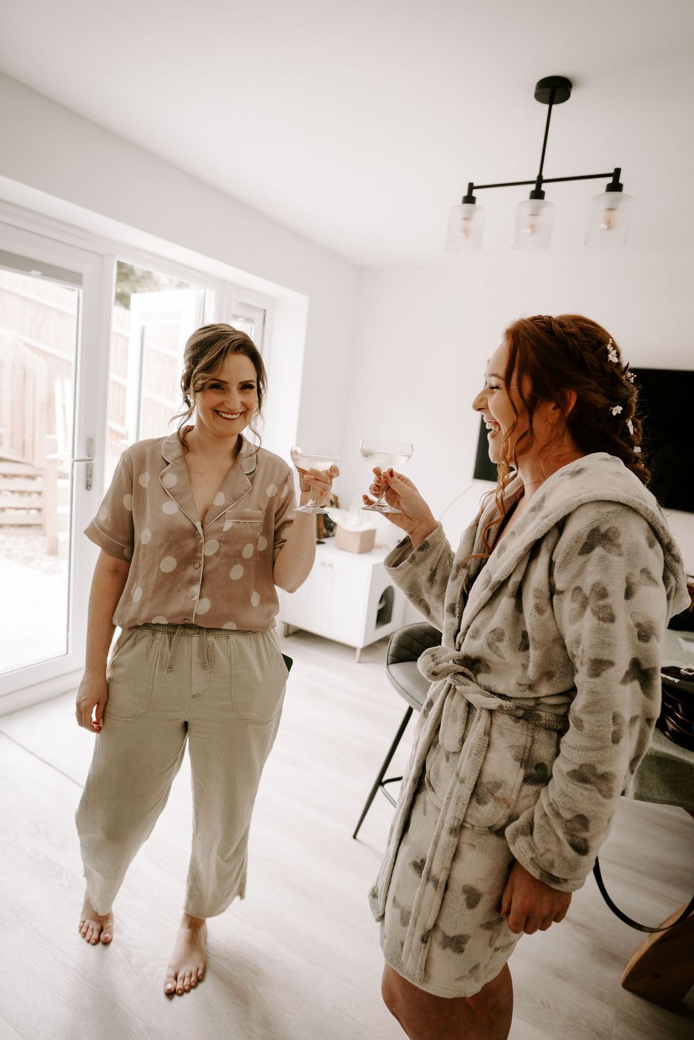 Two women in loungewear toasting with champagne glasses while getting ready for a wedding.