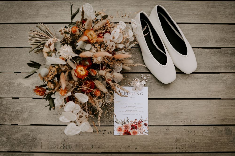 Boho wedding flat lay with dried floral bouquet, white ballet flats, and floral invitation on rustic wood.