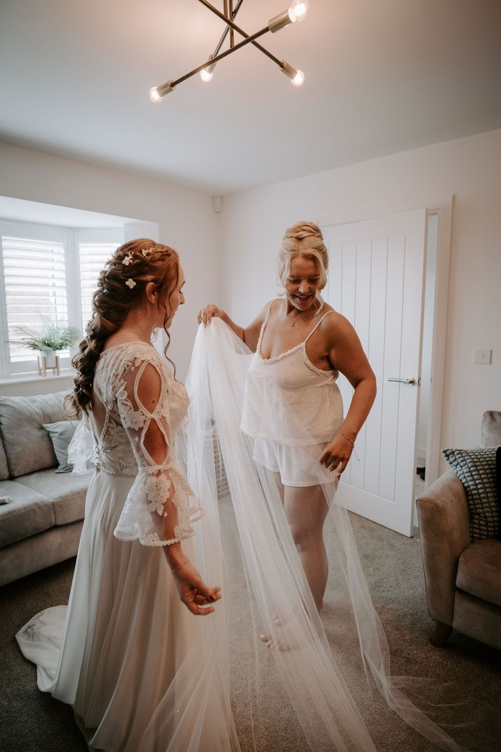 Bride in lace wedding gown gets veil assistance from bridesmaid in white lingerie in a bright living room.