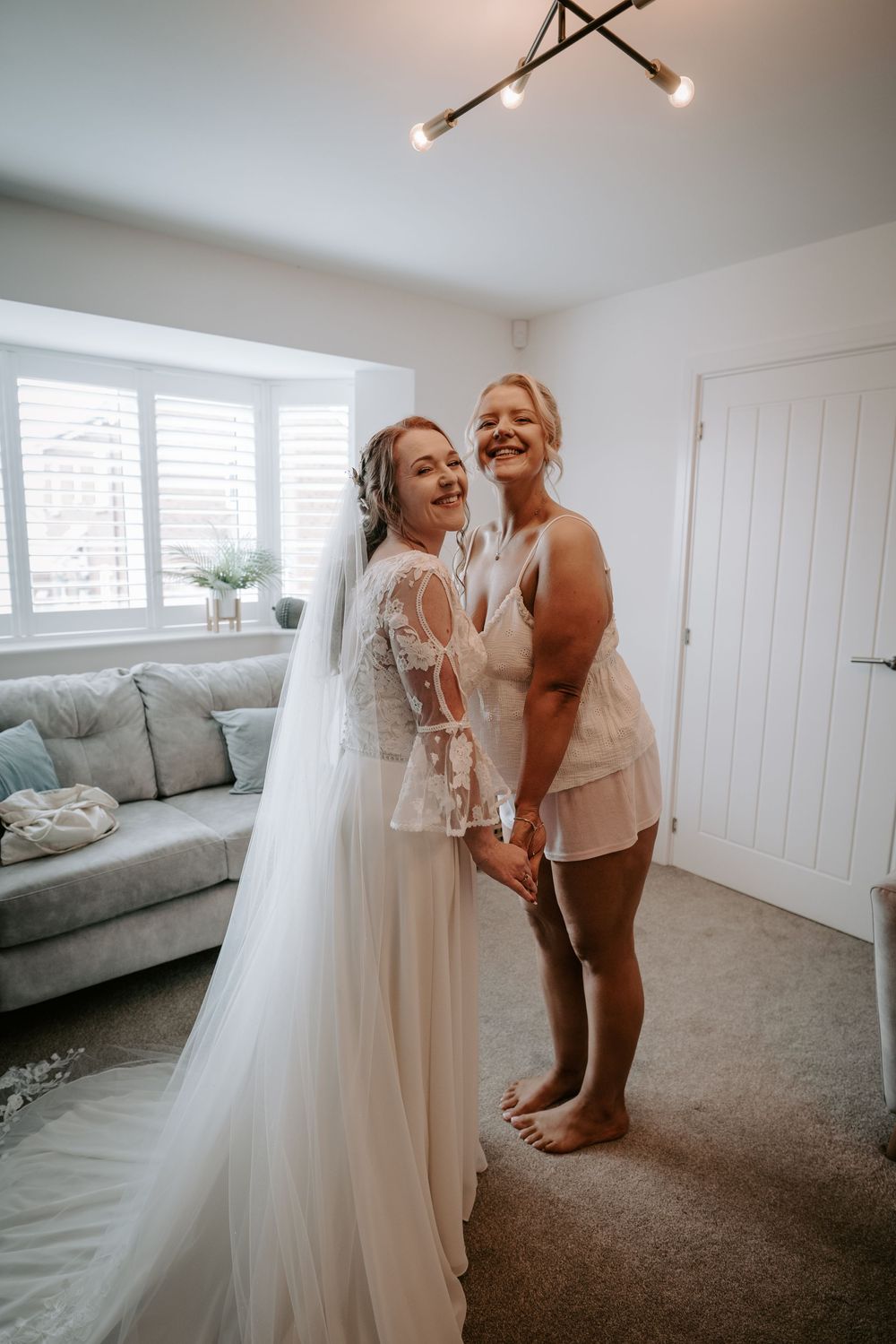 Bride in lace wedding dress and veil holds hands with woman in white lingerie set smiling in bright living room.