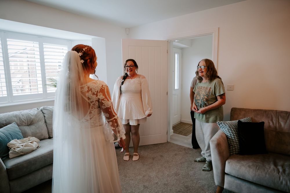 Bride in white gown and veil showing her wedding dress to emotional bridesmaids in a bright living room.