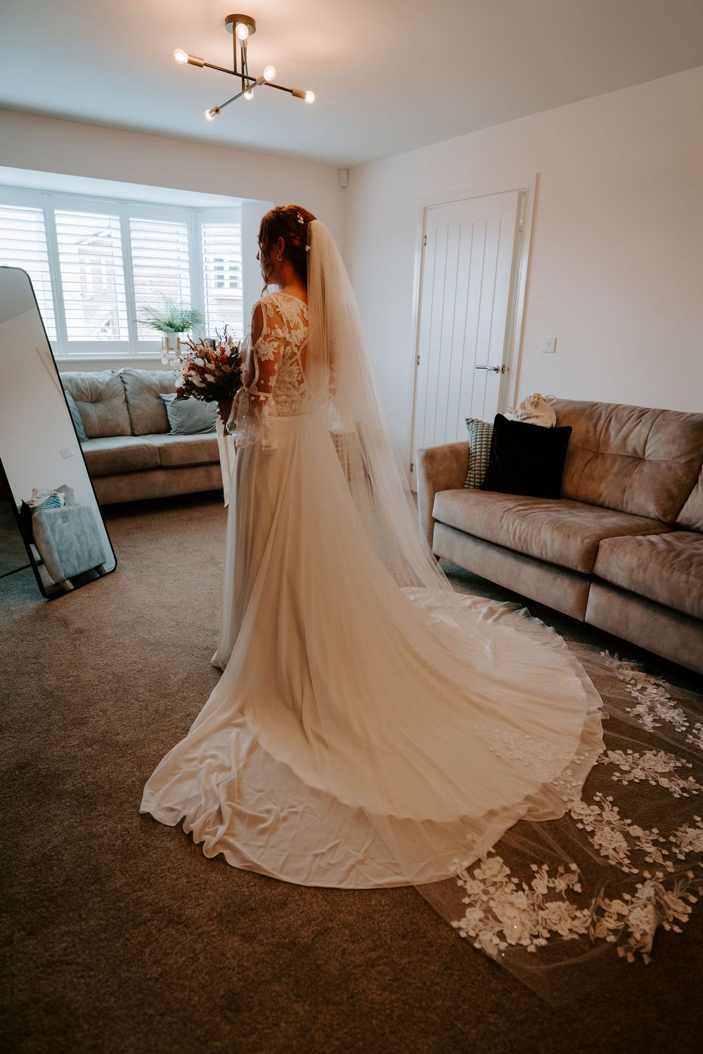 Bride in elegant white lace wedding gown with long veil and floral train stands in a bright living room.
