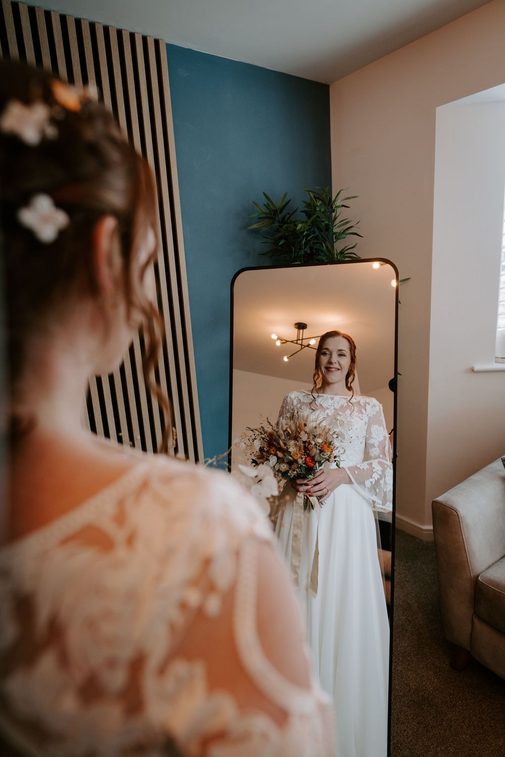 Bride in lace long-sleeve wedding gown holding an autumn bouquet admiring her reflection in a mirror.