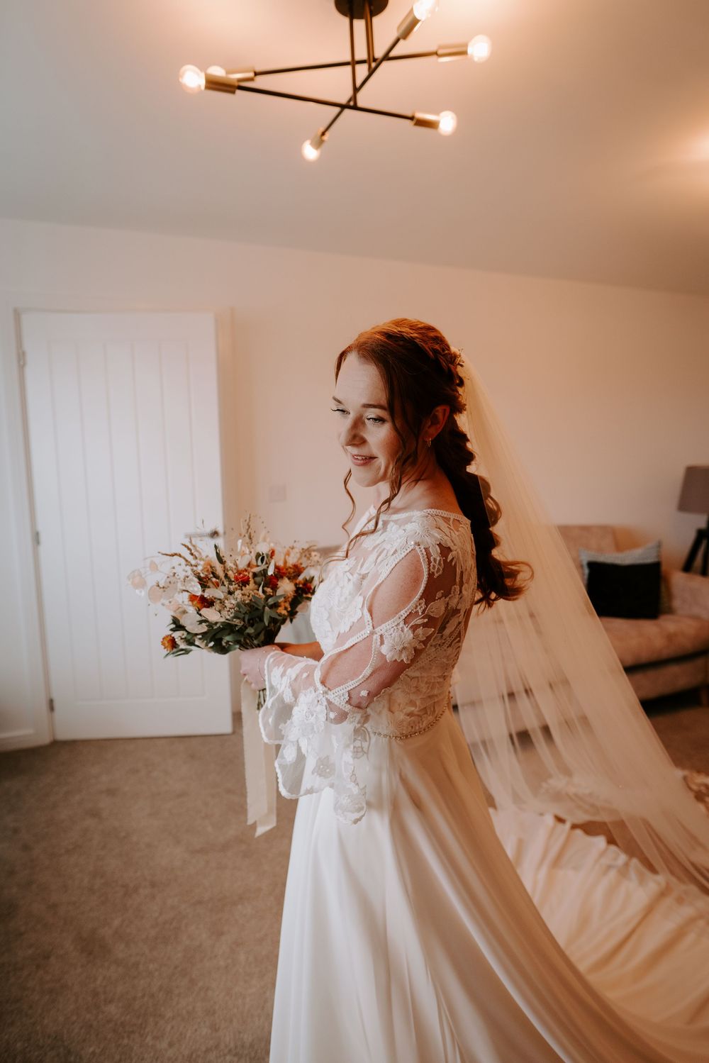 Bride in lace wedding dress with long veil holding a wildflower bouquet while getting ready at home.