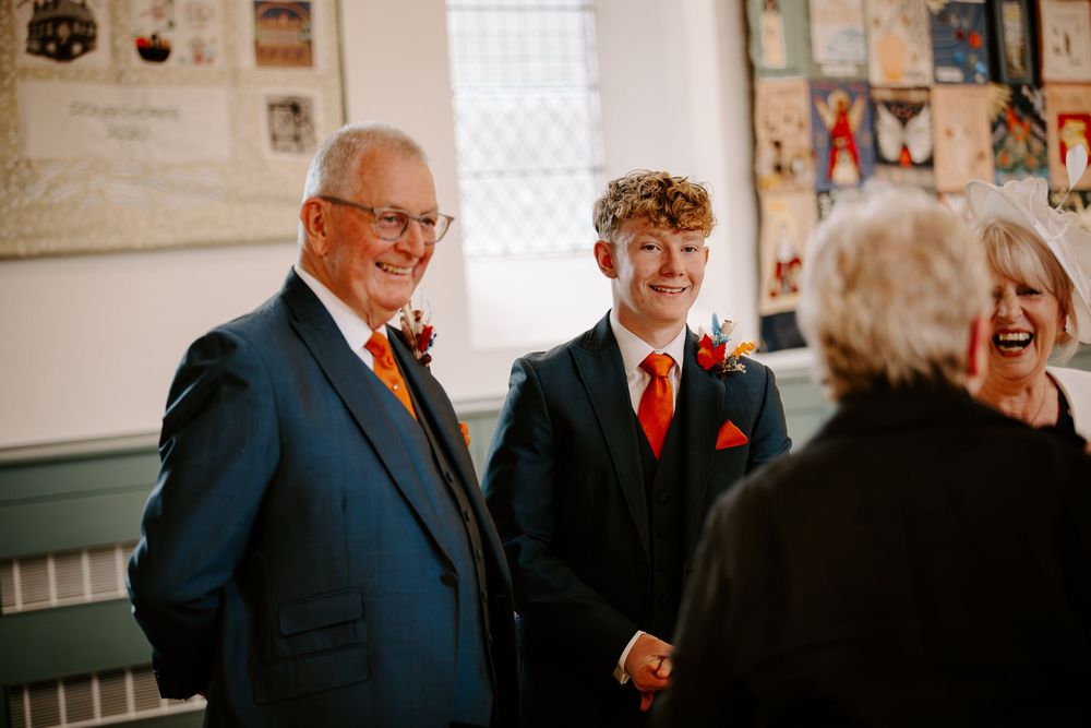 Smiling men in navy suits with orange boutonnieres greet guests at wedding ceremony.