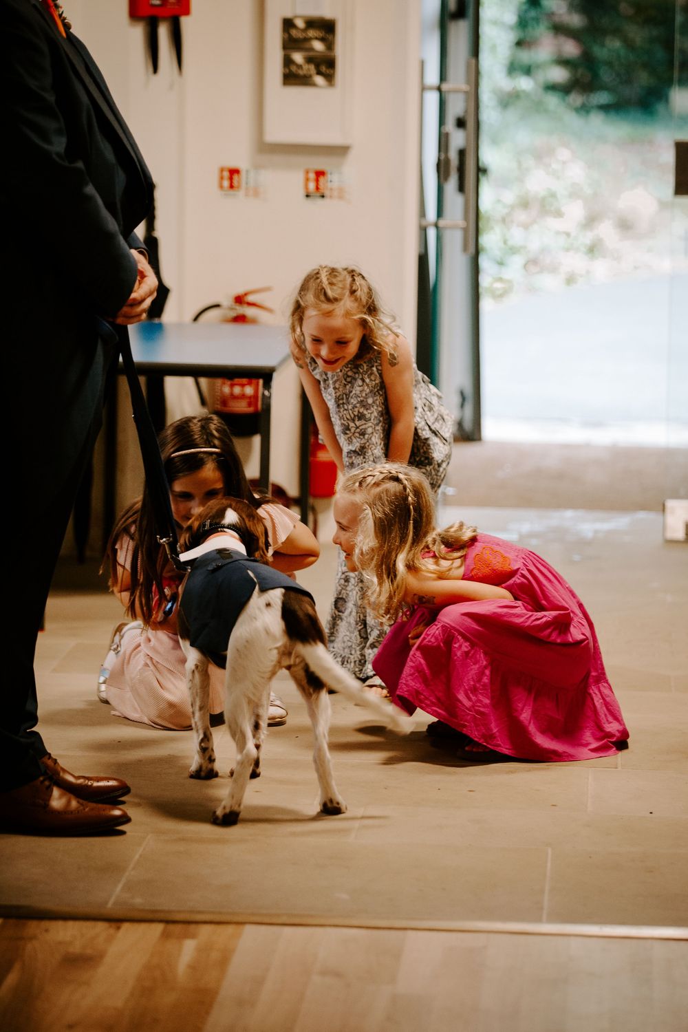 Young flower girls crouching down to pet a small goat wearing a suit jacket at a wedding reception.