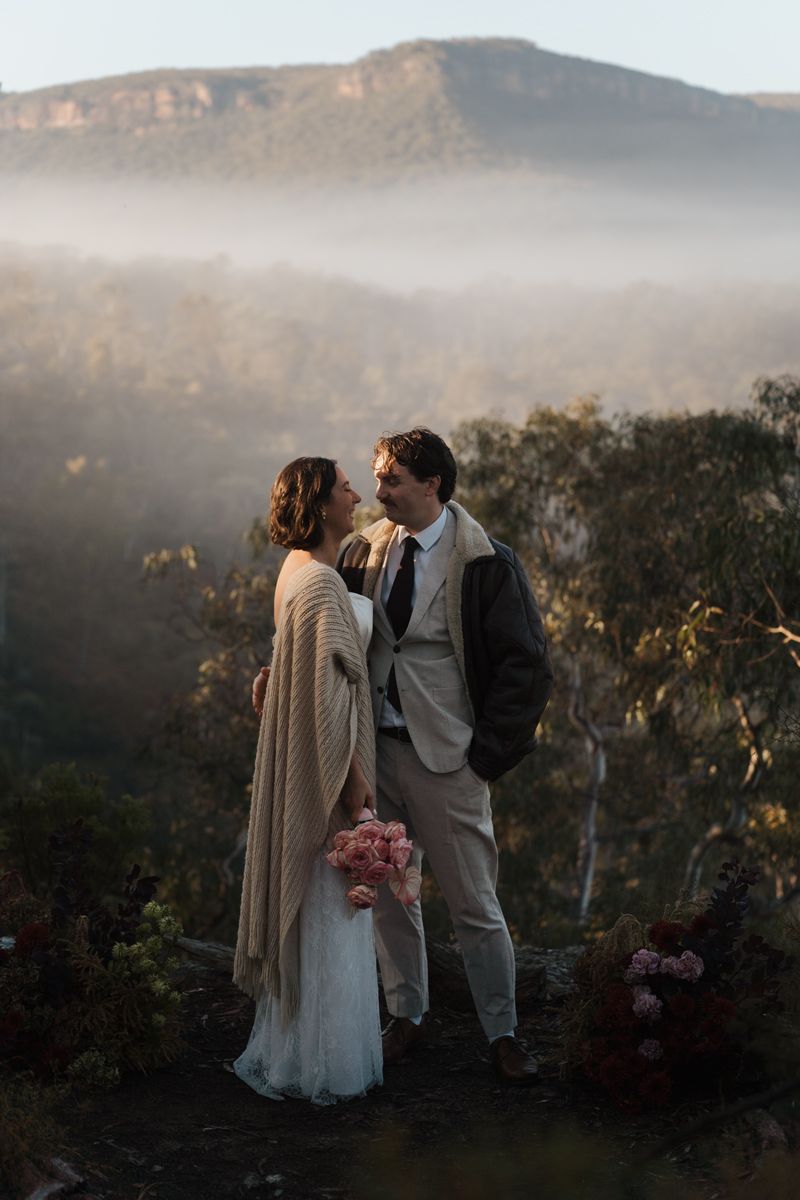 Bride and groom stand on misty mountain overlook, she holds pink bouquet, foggy Blue Mountains valley behind them elopement photoshoot