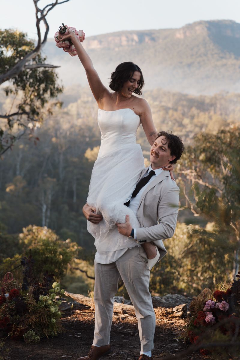 Groom lifting bride in white dress on mountain overlook, celebrating wedding with misty Blue Mountains backdrop elopement photoshoot