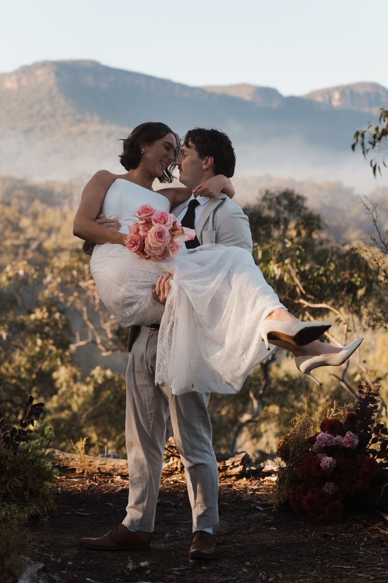 Groom lifts bride in white dress with pink bouquet, sharing a kiss against misty mountain backdrop at outdoor wedding elopement photoshoot