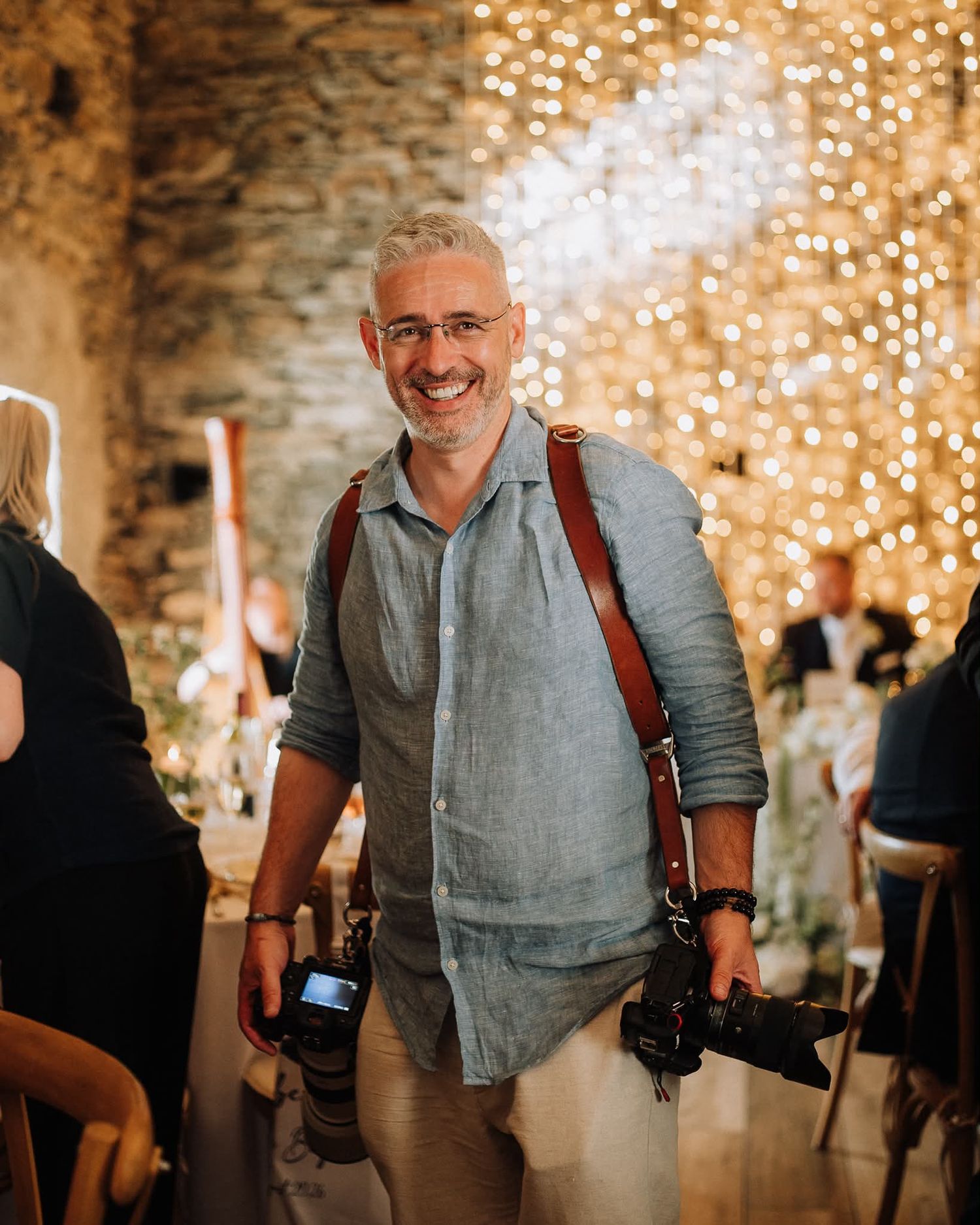 Smiling middle-aged photographer carrying two cameras on leather straps in a warmly lit venue with fairy lights.