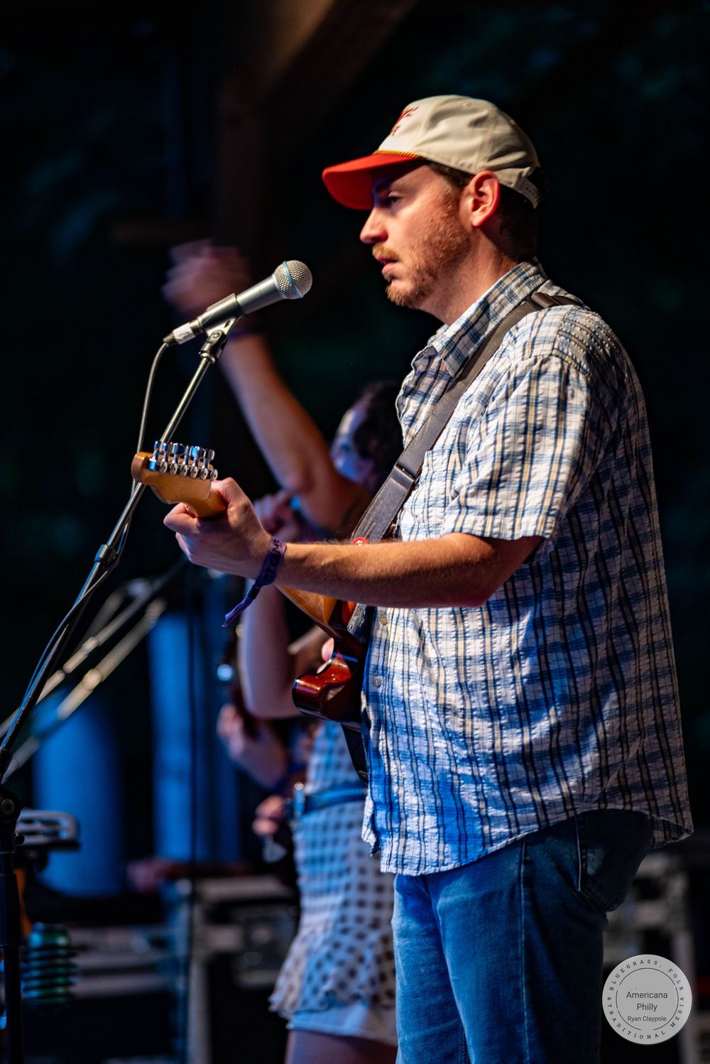 Musician playing guitar and singing into microphone on outdoor stage, wearing plaid shirt and baseball cap.