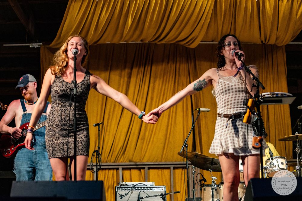 Two women holding hands on stage performing live music with a guitarist visible in the background.