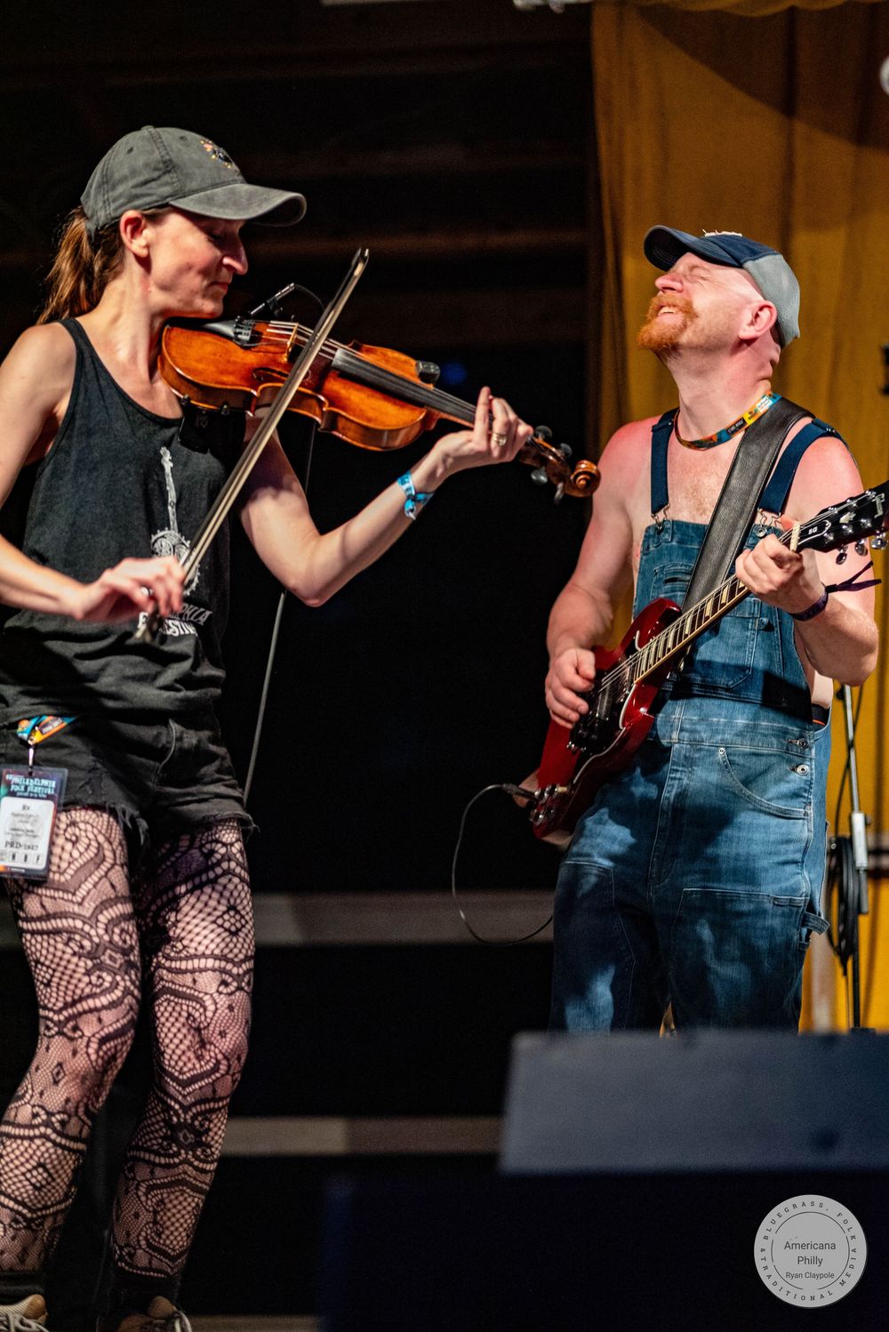 Female violinist and male guitarist in overalls performing together on outdoor festival stage.