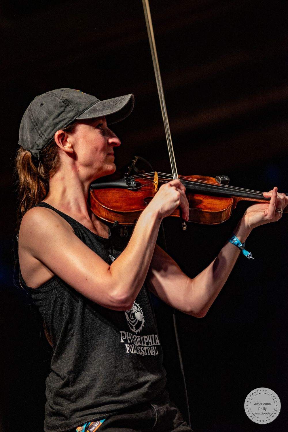 Woman playing violin outdoors at Philadelphia Folk Festival, wearing gray cap and tank top.