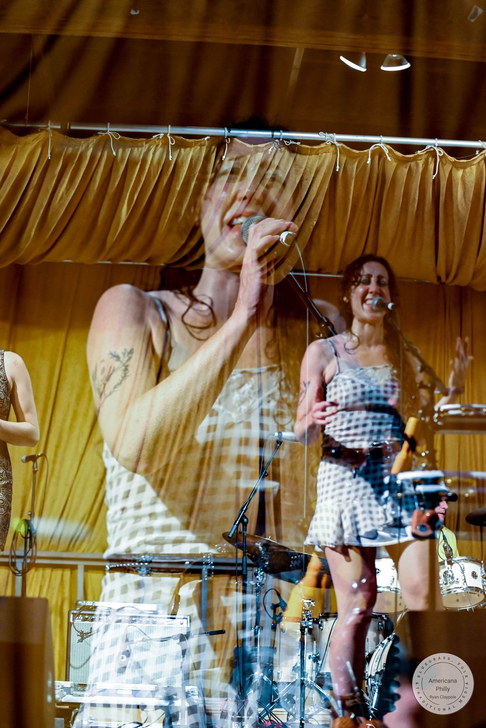 Female performers singing on stage in checkered dresses under golden curtains at a live music event.