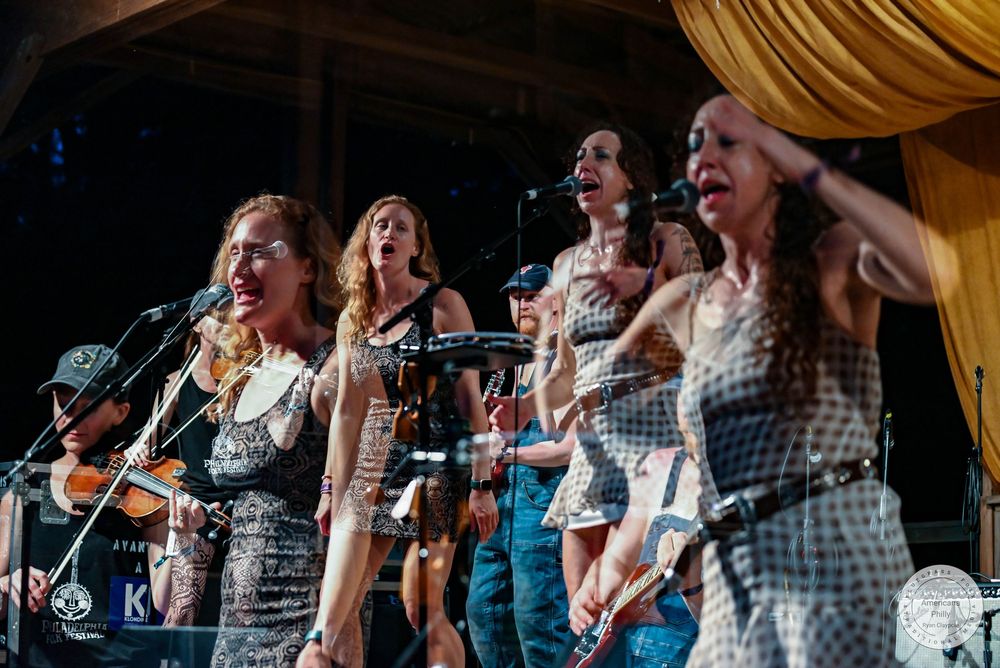 Women in matching dresses perform live on stage, singing and playing instruments at an outdoor music event.